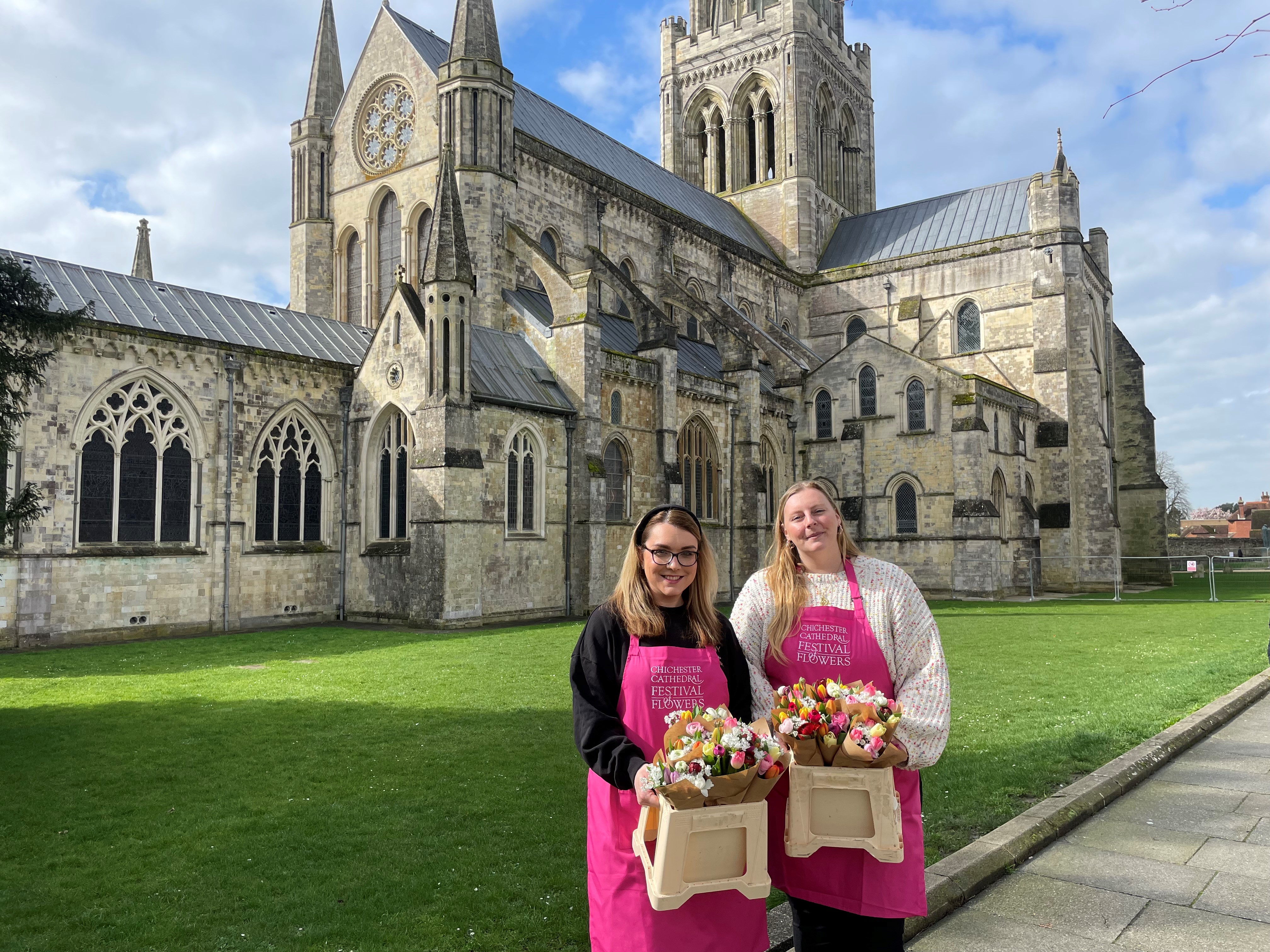 Hannah and Chantal, wearing bright pink aprons, stand in front of the historic grey stone Cathedral, surrounded by green grass. They are holding colourful bunches of flowers.