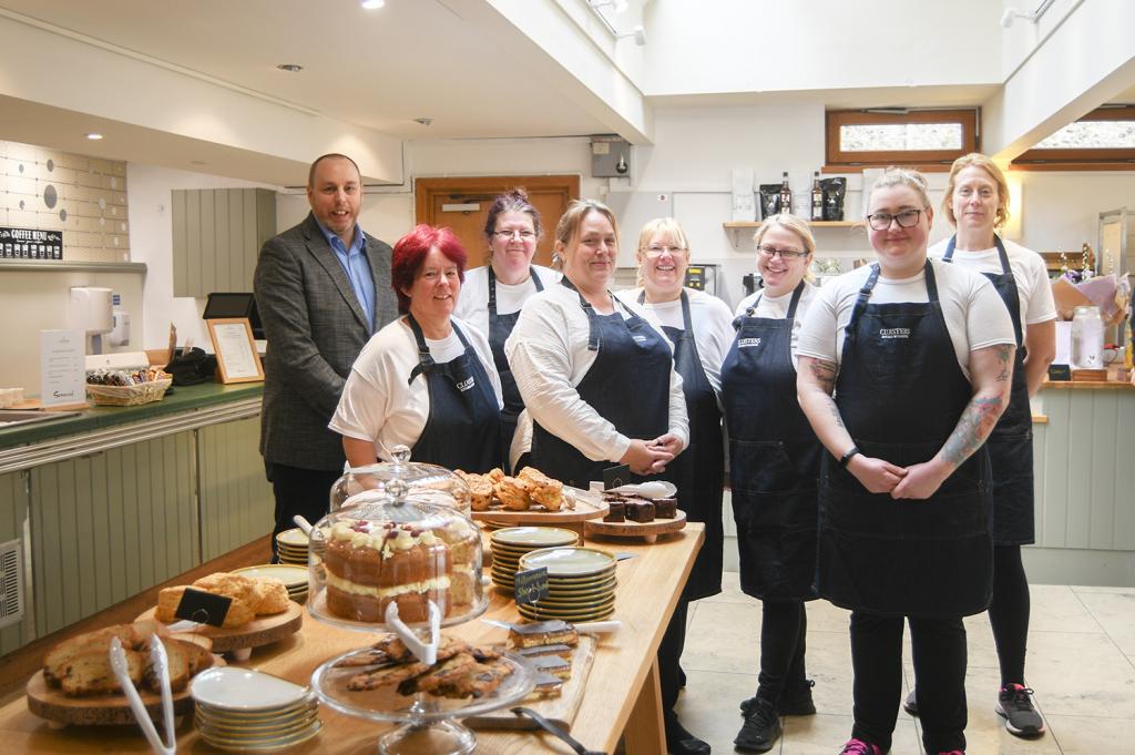 8 people who work in Cloisters Kitchen stand in front of a table of cakes an pastries on opening day.