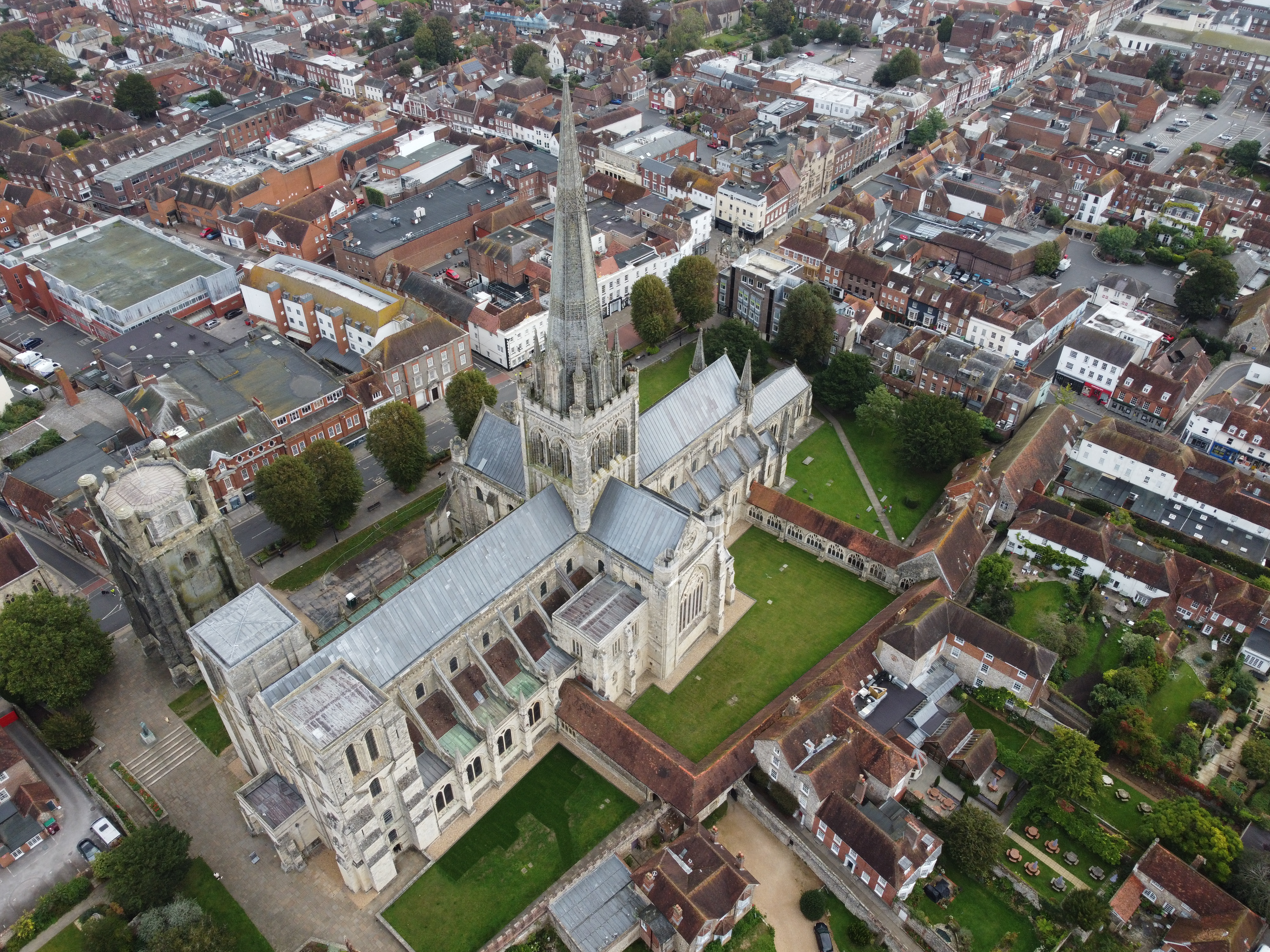 A view of the Chichester Cathedral, covered in grey lead. The historic stone spire sits against a background of buildings - businesses and homes in Chichester