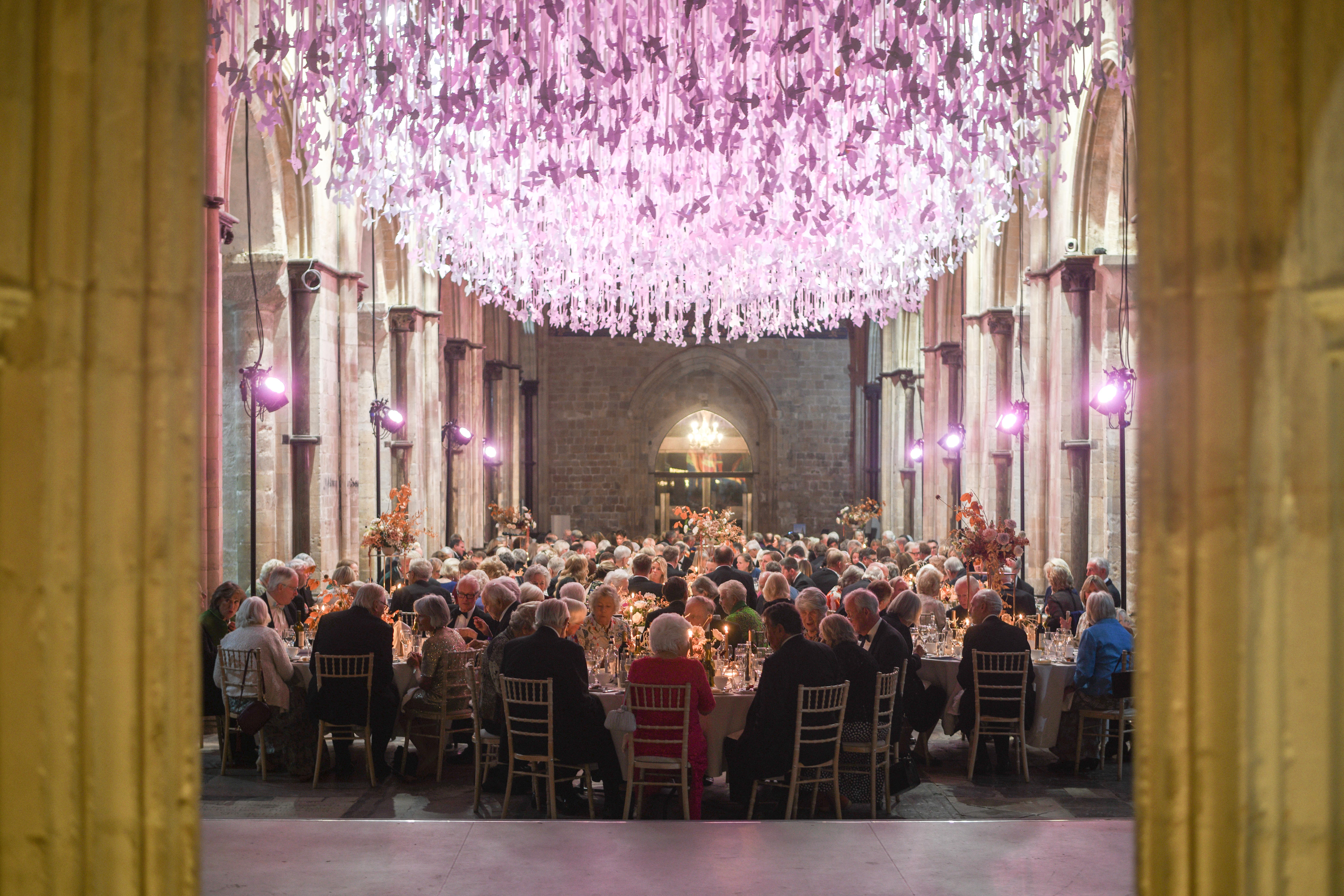 People dressed in formal attire sit around large tables in the Cathedral Nave, under purple Peace Doves