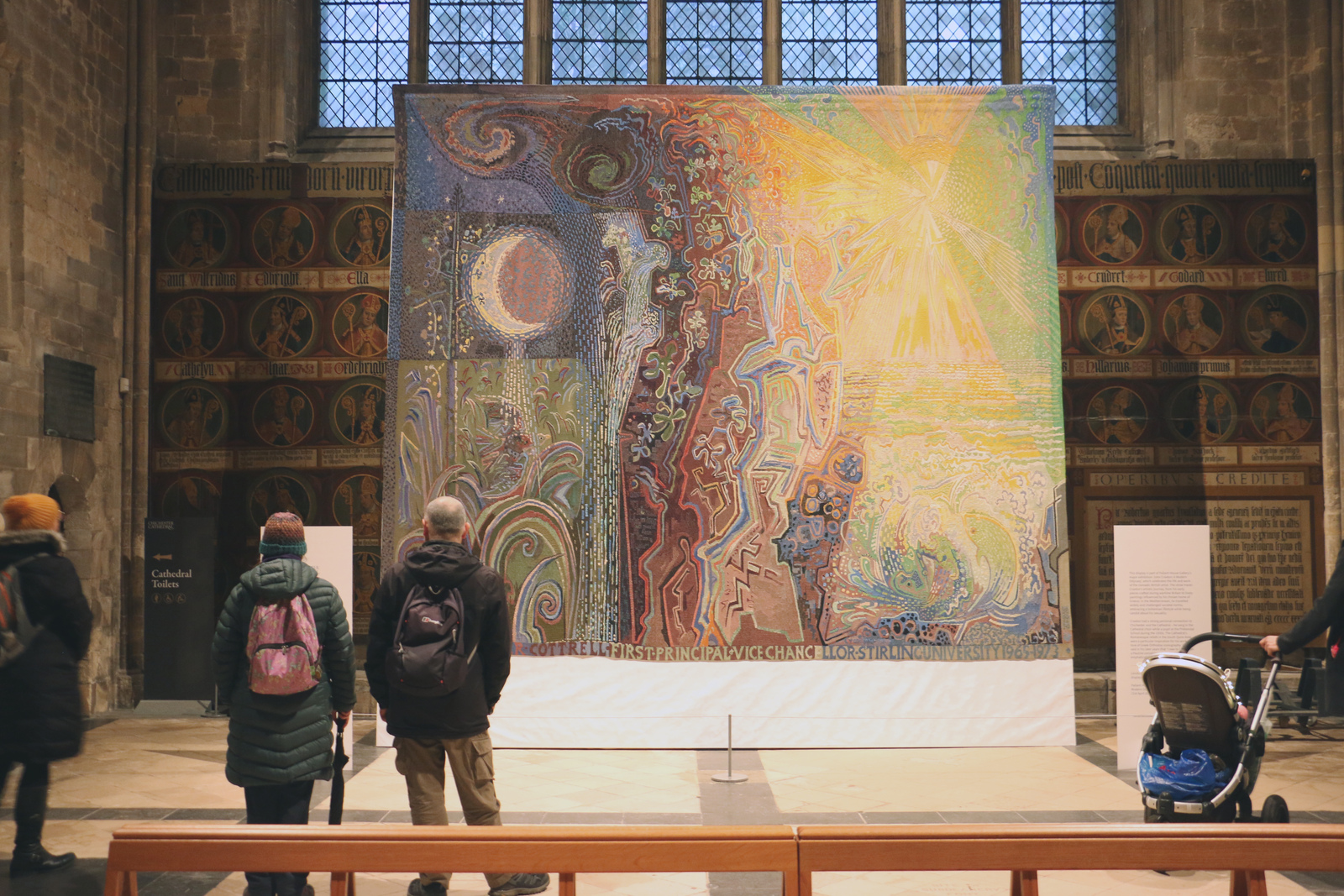 People admiring the large tapestry with their backs to the camera 
