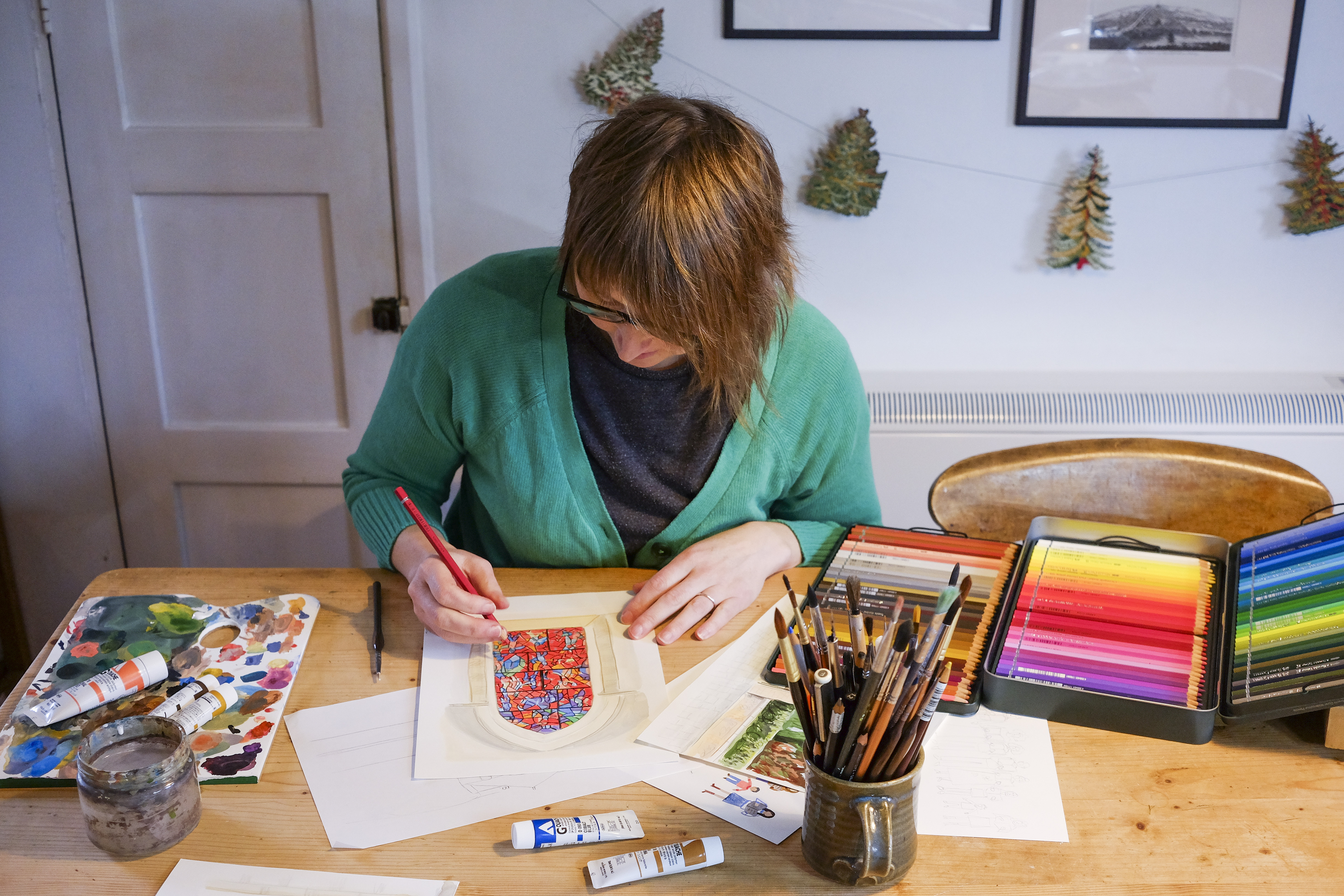 Chloe Robertson, who has brown hair and glasses, colours in her illustration of the Chagall window with a red pencil. The table has pencils, paintbrushes and paints on.
