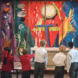 A group of school children face John Piper's colourful tapestry, behind the High Altar. The tapestry has flashes of red, green, blue and yellow. The school children wear red blazers and white shirts.