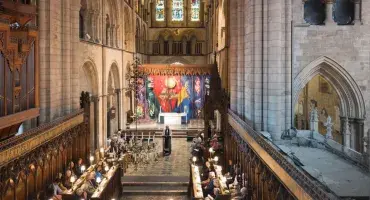 The Cathedral Quire, wooden stalls facing each other with a stone aisle inbetween. The aisle leads to the High Altar - a stone altar which stands in front of a vibrant multi-coloured tapestry by the artist John Piper.