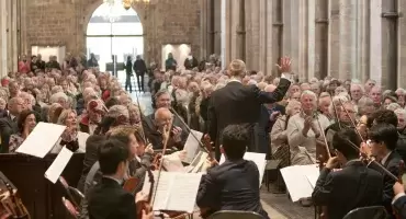 A concert takes place in the Nave, a large group of performers sit with music sheets, people are sitting and clapping in the Nave