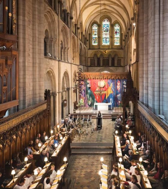 The Cathedral's wooden Quire, with dark wooden stalls facing one another. A paved walkway leads to the High Altar. Behind the stone altar, is the John Piper tapestry, a vibrant and modern artwork with splashes of red, blue, green and yellow.