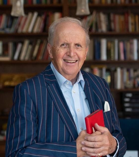 Sir Alexander McCall Smith stands in a library, holding one of his books
