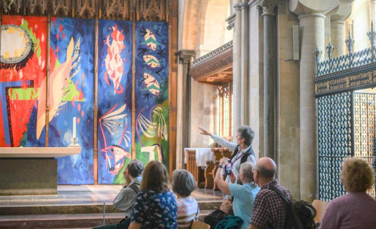 Visitors seated in the Cathedral watch as a Guide explains a colourful contemporary artwork displayed behind the altar, framed by historic stone columns and carved woodwork.