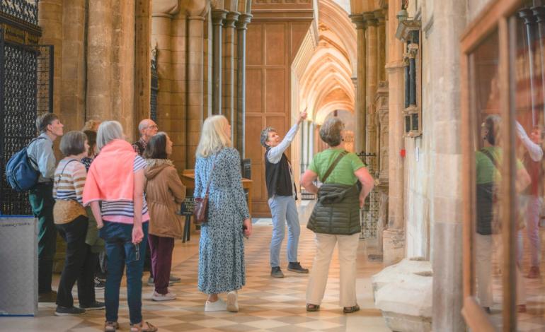 A group of visitors follows a Cathedral Guide through the nave aisle, looking up at architectural features and historic memorials during a guided tour of the Cathedral.