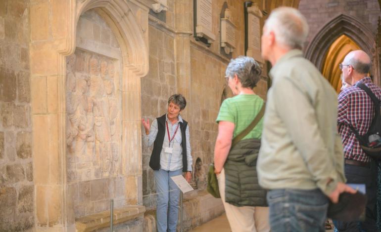A Cathedral Guide speaks to visitors beside a carved medieval stone relief set within an arched recess in the Cathedral wall, while a small group gathers to listen in the nave aisle.