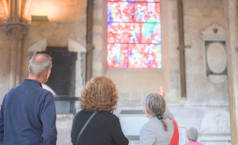 Visitors pause to admire and discuss a colourful stained-glass window by Marc Chagall, surrounded by historic stone walls, memorials and monuments.