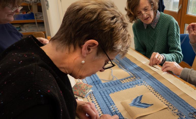 A woman embroiders blue thread for a kneeler