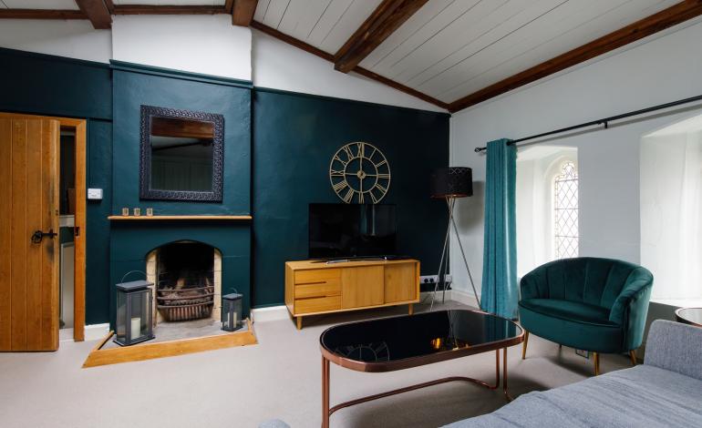 A lounge with grey sofas and dark teal walls, a flat screen TV, fireplace, coffee table and gold clock on the wall