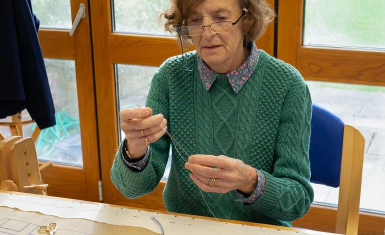 A woman sits in front of blue thread weaving through a needle, creating an embroidered piece