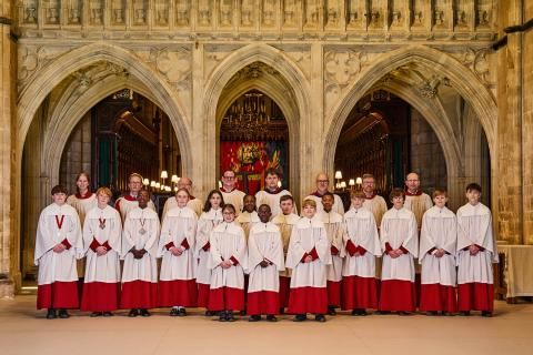 The full Cathedral Choir pose, lined up in three rows, in front of the historic stone-arched Arundel Screen. The Choir wear red and white cassocks, with Choristers stood at the front and Lay Vicars at the back.