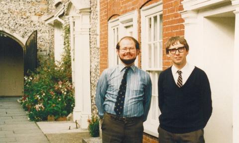 Two white men in late 80s clothing standing in front of a red brick building