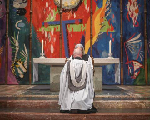 Clergy member in white and black vestments kneeling at the High Altar in Chichester Cathedral, with John Piper’s vibrant High Altar Tapestry filling the background in bold reds, blues and greens