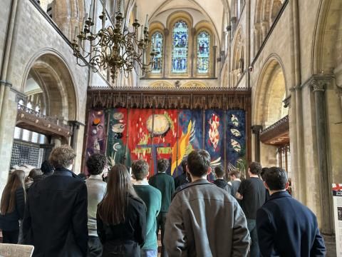 Inside Chichester Cathedral, a group of people stand in front of a large, brightly coloured tapestry behind the altar. The tapestry includes abstract shapes in red, blue and yellow. Above it, tall arched windows and an ornate chandelier hang beneath the vaulted stone ceiling.