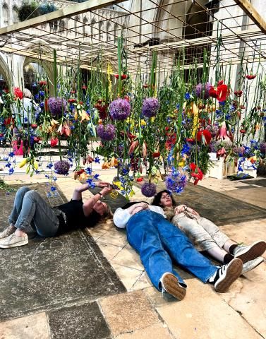 Inside Chichester Cathedral, three Plumpton College students lie on the stone floor beneath a suspended floral installation. Dozens of colourful flowers—such as purple alliums, blue delphiniums, red and yellow blooms—hang down from a rectangular wooden grid above them. The students are reaching up and interacting with the hanging stems while the cathedral’s stone arches rise in the background.