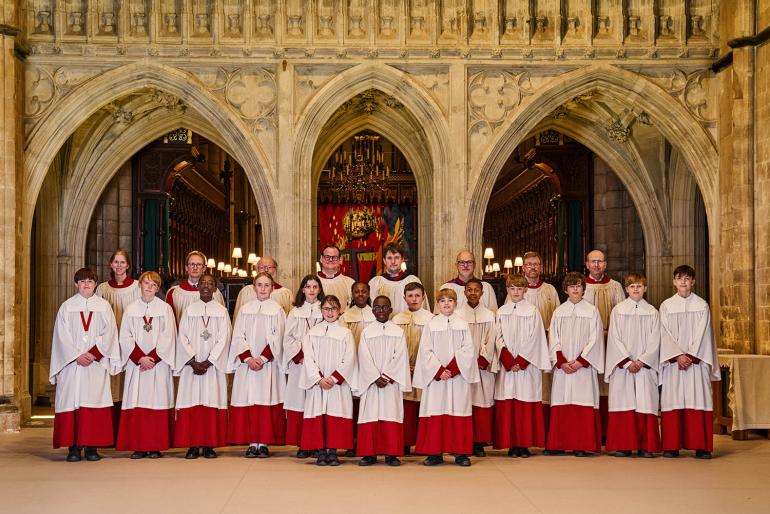The full Cathedral Choir pose, lined up in three rows, in front of the historic stone-arched Arundel Screen. The Choir wear red and white cassocks, with Choristers stood at the front and Lay Vicars at the back.