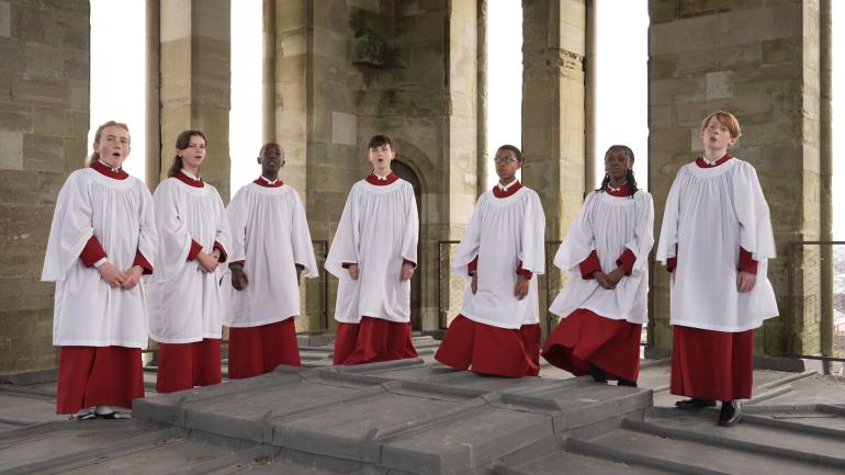 Seven Choristers stand within the Cathedral's Lantern, surrounded by stone arches, and wearing red and white cassocks.
