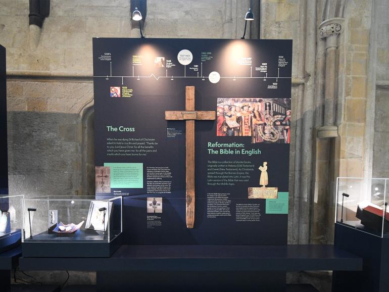 A wooden cross sitting amongst a busy exhibition, containing writing and images. The cross has a pointed bottom edge, which is worn - perhaps where it has been fixed into the ground.