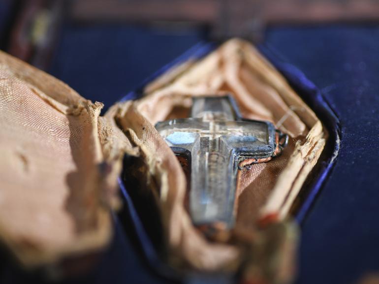 A glass cross, sitting within a beige, warn diamond-shape pocket and surrounded by rich purple fabric.