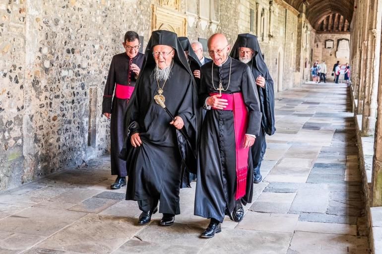 His All Holiness Ecumenical Patriarch Bartholomew  walks alongside The Bishop of Chichester in the Cathedral's stone Cloisters. They are followed by the Dean of Chichester and other clergy.  The Patriarch wears traditional black robes. The Bishop and Dean also wear black robes, but with magenta sashes.