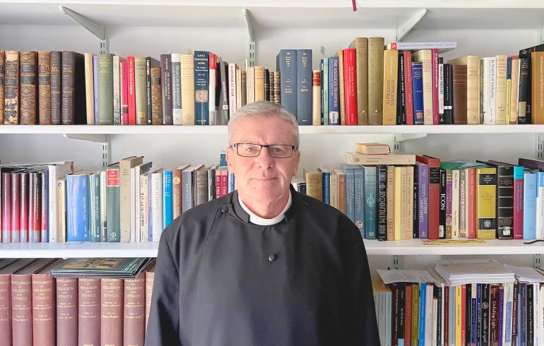 Reverend Dr Earl Collins, a man man wearing clerical attire, including a black cassock with a white clerical collar, stands in front of a bookshelf filled with a variety of academic and religious books. He has short grey hair, glasses, and a calm expression.
