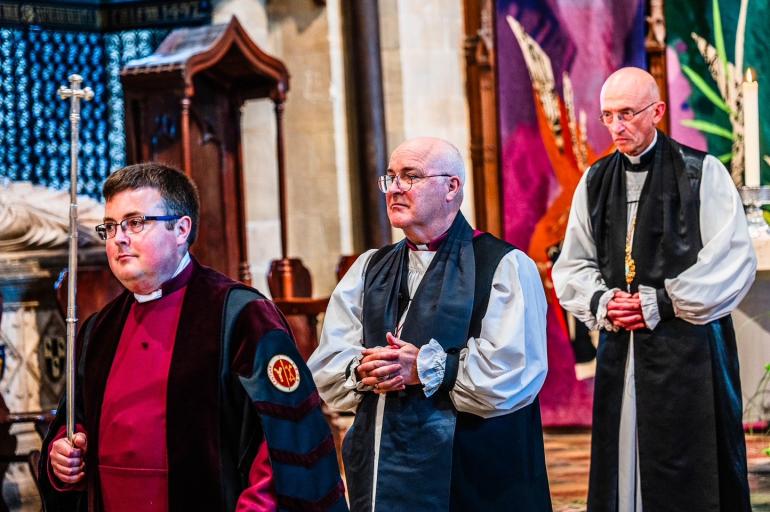 The Archbishop of York and Bishop of Chichester process through the Cathedral during a service, led by the Cathedral's Head Verger. The Head Verger carries a long silver verge and wears a vibrant burgundy cassock.