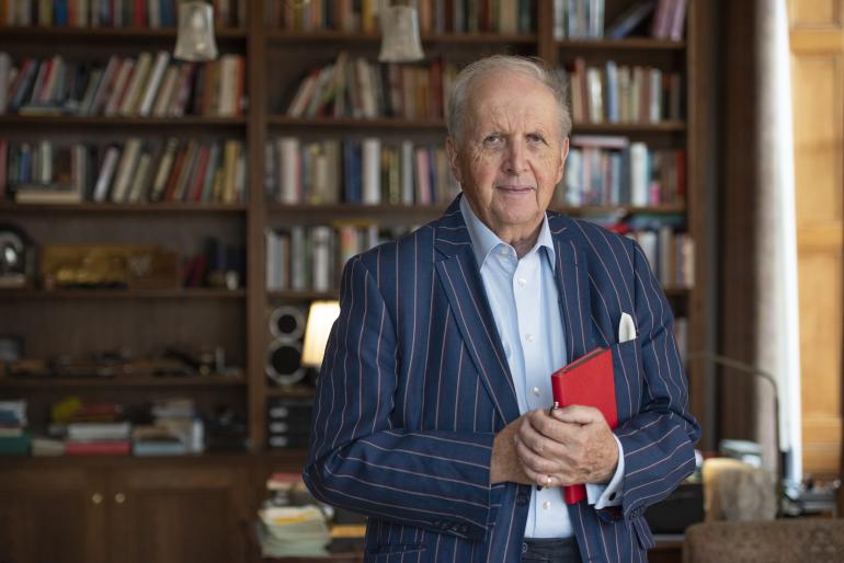 Sir Alexander McCall Smith holds a red book, wearing a pinstripe navy blazer and blue tie. Behind him is a full, wooden bookshelf.