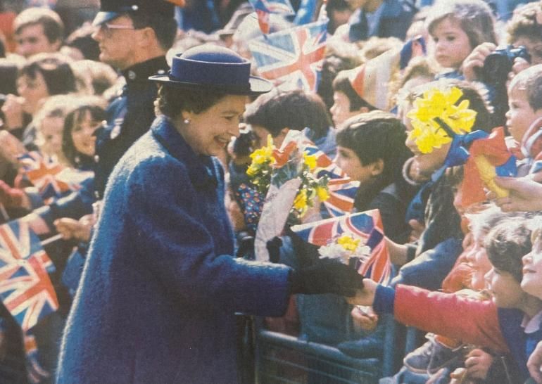 A well dressed woman is blue shaking the hand of a small child who's part of a big crowd many holding flowers or Union Jack flags.