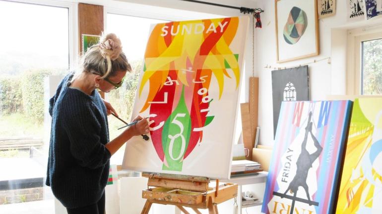 Carter painting in her studio, East Sussex (2025). The artist paints onto a canvas with bright colours of orange, yellow, green and red. She has light hair and dark clothing. Her bright studio, with view of a nearby green space, has pictures on walls from floor to ceiling. 