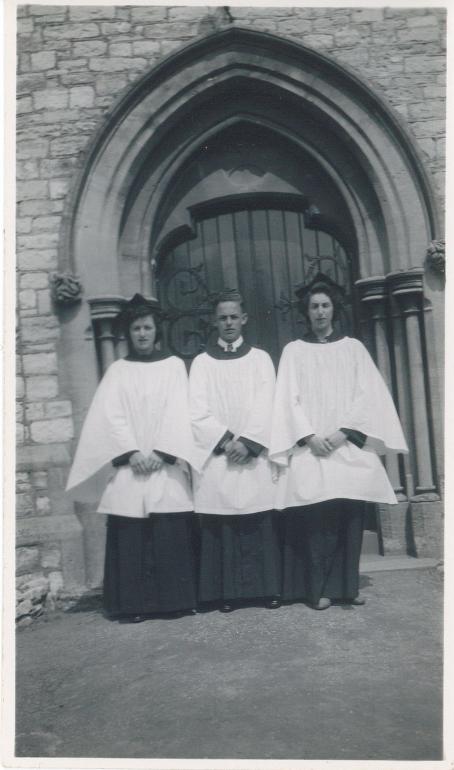 B&W wtih two women and a man wearing choir robes looking to camera in front of cathedral door