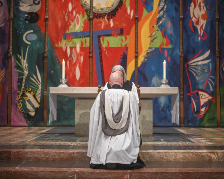 Clergy member in white and black vestments kneeling at the High Altar in Chichester Cathedral, with John Piper’s vibrant High Altar Tapestry filling the background in bold reds, blues and greens