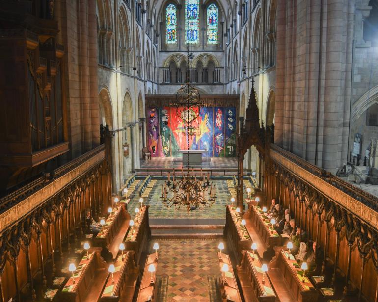 A wide view down the choir stalls of Chichester Cathedral toward the High Altar, with John Piper’s High Altar Tapestry brightly illuminated at the far end beneath the cathedral’s stained-glass windows.