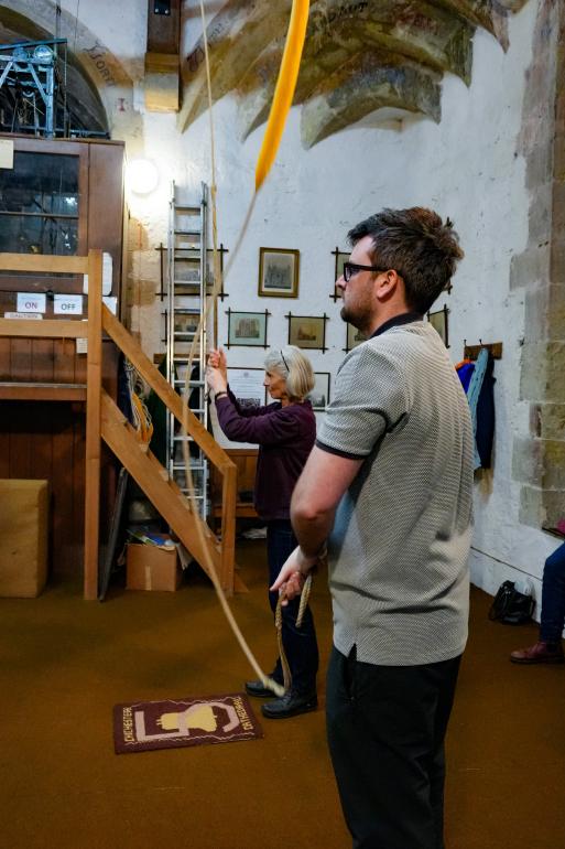 Two people stand inside the bell‑ringing chamber holding bell ropes, one in the foreground and one farther back. Behind them are wooden stairs leading to an upper platform, a ladder, framed artwork, and stone walls with arched features. The ropes extend upward toward the ceiling.