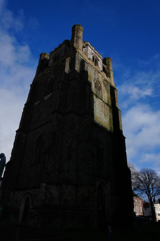 A tall, stone bell tower of Chichester Cathedral photographed from a low angle, with most of the tower in deep shadow. Sunlight highlights the upper sections of the weathered masonry. The tower rises against a bright blue sky with scattered clouds, and leafless trees and nearby buildings are visible at the edge of the frame.