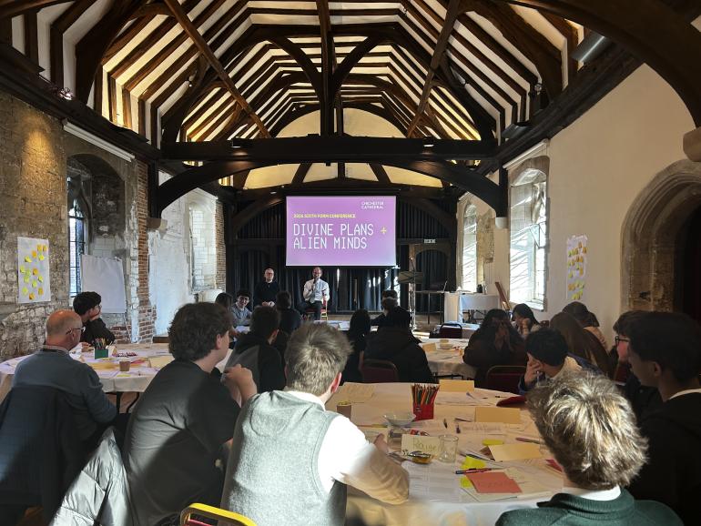 Inside Vicars hall at Chichester Cathedral, people sit around large tables covered with papers, pens and workshop materials. At the front of the room, two speakers stand beneath a projected presentation slide titled “DIVINE PLANS + ALIEN MINDS.” The space has exposed timber beams, stone walls and tall arched windows letting in natural light.