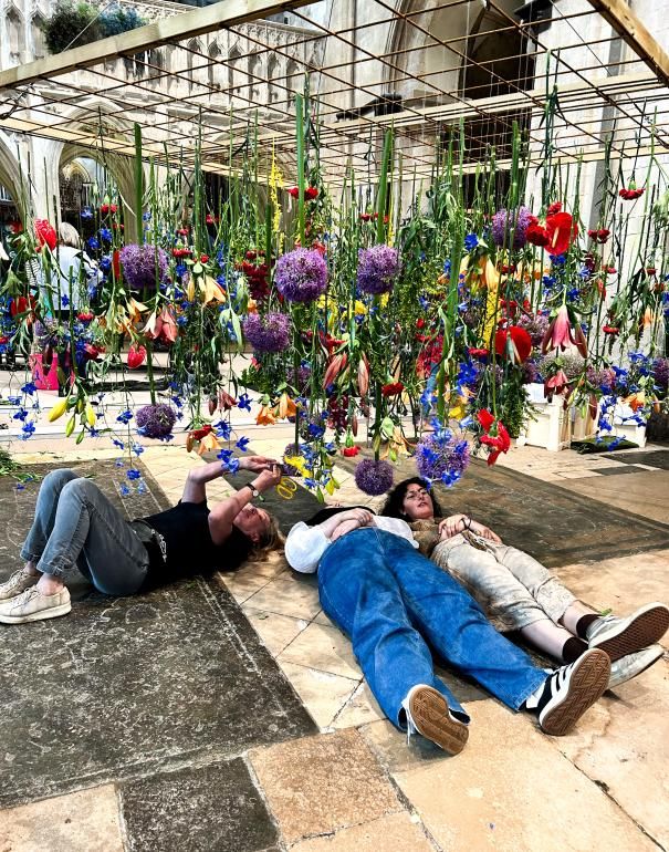 Inside Chichester Cathedral, three Plumpton College students lie on the stone floor beneath a suspended floral installation. Dozens of colourful flowers—such as purple alliums, blue delphiniums, red and yellow blooms—hang down from a rectangular wooden grid above them. The students are reaching up and interacting with the hanging stems while the cathedral’s stone arches rise in the background.