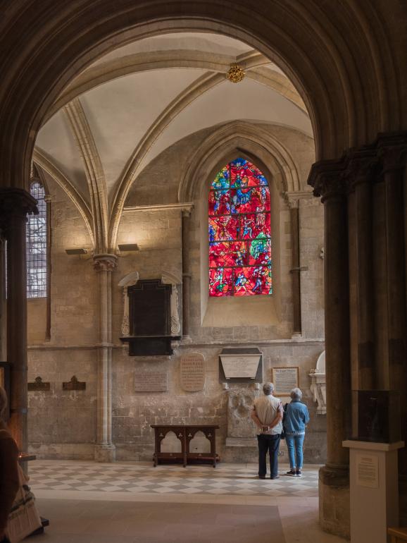 A tall, arched stained-glass window by Marc Chagall glows with vivid reds, blues, and greens, set into the stone wall of Chichester Cathedral. The window is seen through medieval arches and columns beneath a ribbed vaulted ceiling, with memorial plaques on the wall below and two visitors standing quietly, looking up at the artwork.