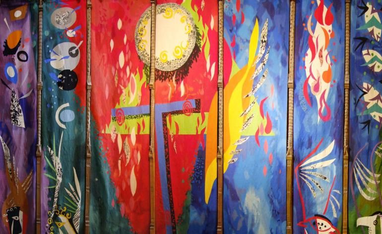 The Piper Tapestry showing tall vertical panels filled with bold reds, blues, and greens, featuring abstract birds, flames, leaves, and a glowing circular sun‑like form.