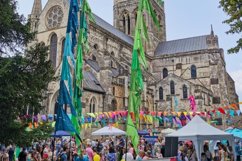 Crowds gathered in front of Chichester Cathedral. Multicoloured bunting is hung from the Cathedral and festival flags in blue and green dominate the image.