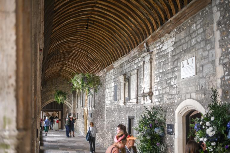 Visitors walk through the cloisters at Chichester Cathedral, beneath a timber-vaulted ceiling and alongside stone walls lined with memorial plaques, as large floral installations and hanging greenery decorate the corridor during the Festival of Flowers 2024
