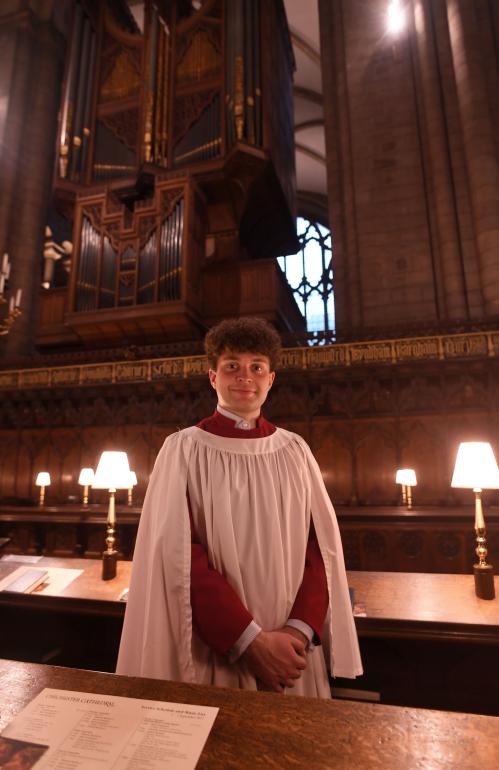 Organ scholar, Benedict Tanner, standing in choir stalls beneath a large wooden pipe organ inside a cathedral, wearing a red cassock and white surplice, with warm lamps and carved wood panelling visible in the background.
