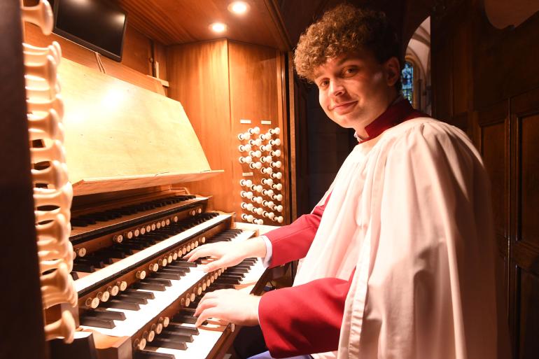 Organ scholar Benedict Tanner playing a multi-manual pipe organ console inside a cathedral, wearing a red cassock and white surplice, with rows of stop knobs and warm wooden panelling lit by overhead lights.